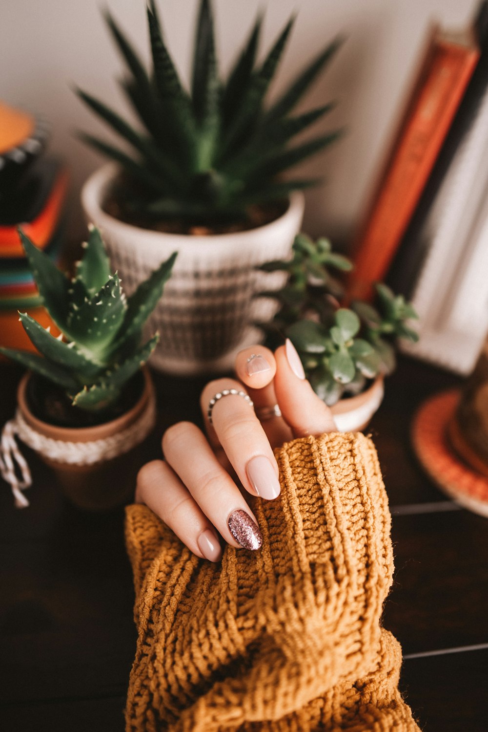 Nails finished in soft brown against a mustard knit sleeve, houseplants behind