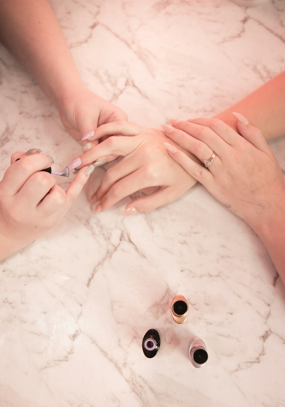 A technician painting a client's nails at a pink marble table, polish bottles beside them